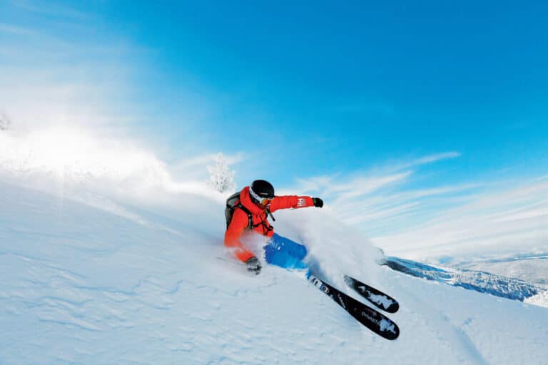 En skiløber i rød jakke og blå bukser tager et skarpt sving på en sneklædt skråning og sparker en spray af sne op. Himlen er klar og blå, og bjergrigt terræn er synligt i baggrunden.