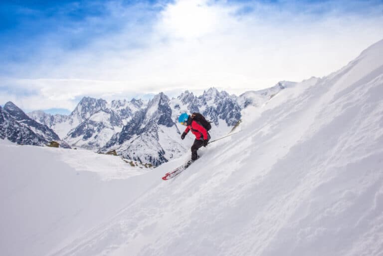 En skiløber iført blå hjelm og rød jakke går ned ad en stejl, sneklædt bjergside. Scenen er sat på baggrund af barske, snedækkede tinder under en lys, overskyet himmel.