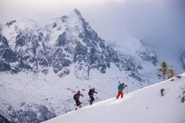 Tre skiløbere vandrer op ad en sneklædt bjergskråning med ski på rygsækken. Den store bjergkæde og forrevne tinder er synlige i baggrunden under en overskyet himmel.