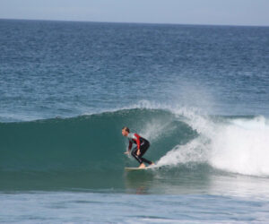 En surfer rider på en bølge på en dag med klart hav. Personen er iført en sort og rød våddragt og balancerer dygtigt på et surfbræt, mens bølgen krøller sig omkring dem. Horisonten er synlig i baggrunden under en blå himmel.