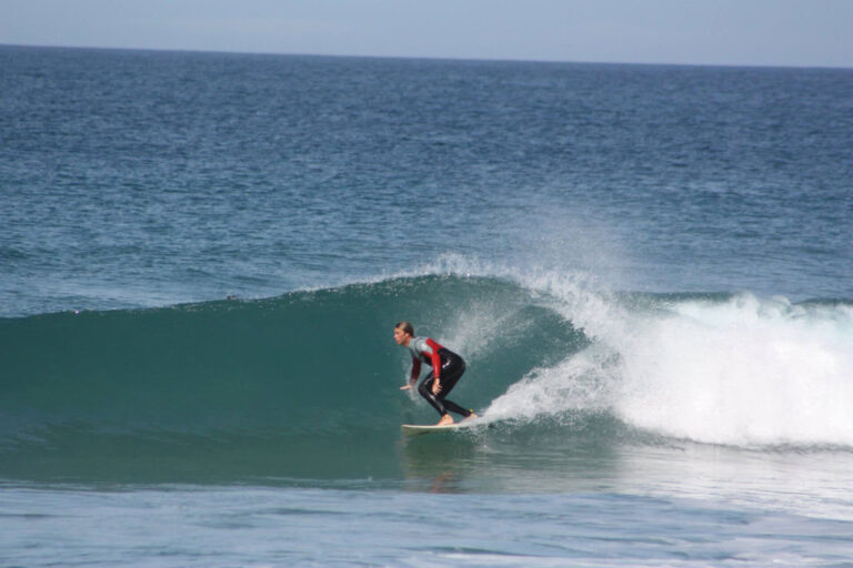 En surfer rider på en bølge på en dag med klart hav. Personen er iført en sort og rød våddragt og balancerer dygtigt på et surfbræt, mens bølgen krøller sig omkring dem. Horisonten er synlig i baggrunden under en blå himmel.