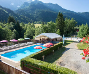 En naturskøn udsigt over et resort med en udendørs pool omgivet af liggestole og parasoller. Et hvidt telt er i nærheden, med en kulisse af frodige grønne træer og majestætiske bjerge under en klar blå himmel.