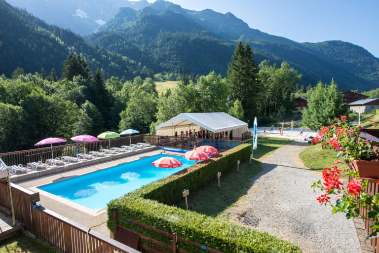 En naturskøn udsigt over et resort med en udendørs pool omgivet af liggestole og parasoller. Et hvidt telt er i nærheden, med en kulisse af frodige grønne træer og majestætiske bjerge under en klar blå himmel.