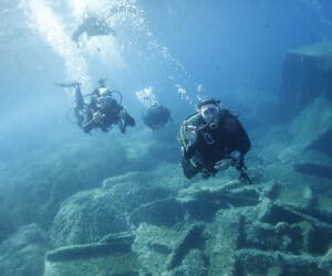 Fire dykkere udforsker et undervandslandskab med klippeformationer. Klart blåt vand omgiver dem, og bobler stiger, mens de svømmer. Scenen er rolig og solrig, hvilket fremhæver skønheden i undervandsmiljøet.