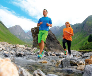 En mand og en kvinde jogger gennem en stenet bjergstrøm. Manden er iført blå skjorte og grønne shorts, og kvinden er i orange skjorte og sorte bukser. Baggrunden har grønne bakker og en overskyet himmel.