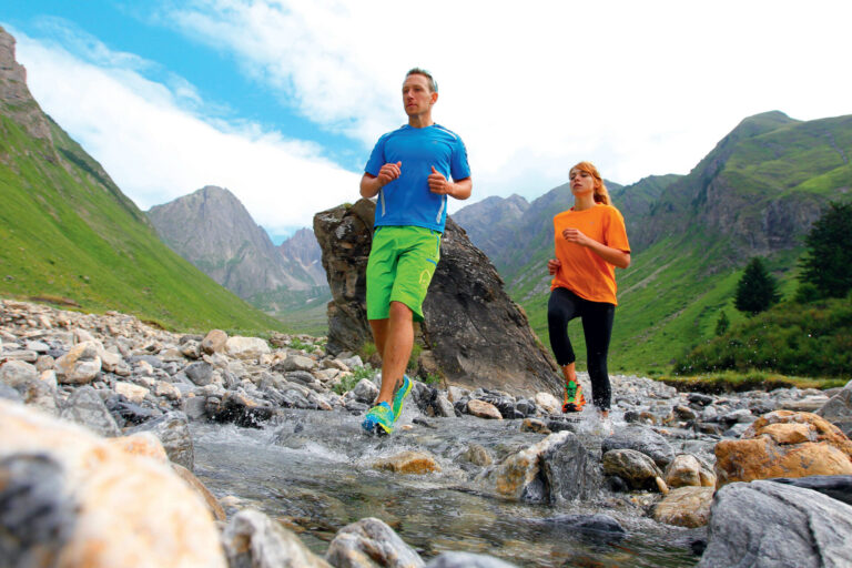 En mand og en kvinde jogger gennem en stenet bjergstrøm. Manden er iført blå skjorte og grønne shorts, og kvinden er i orange skjorte og sorte bukser. Baggrunden har grønne bakker og en overskyet himmel.