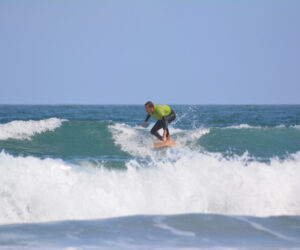 En person i en grøn skjorte og våddragt surfer på en bølge i havet. Bølgen er mellemstor med hvidt skum, og himlen ovenover er klar og blå.