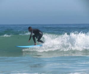 En surfer i en sort våddragt rider på en lille bølge på et blåt surfbræt i klart havvand under en klar himmel.