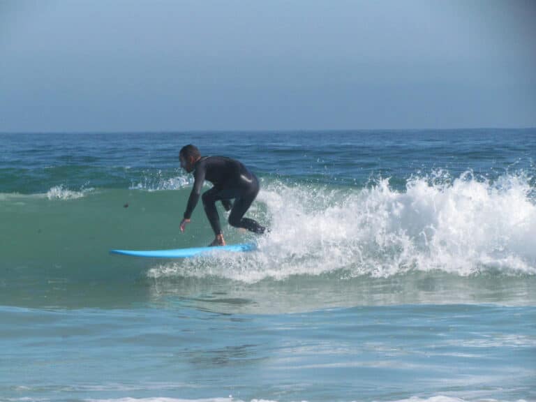 En surfer i en sort våddragt rider på en lille bølge på et blåt surfbræt i klart havvand under en klar himmel.