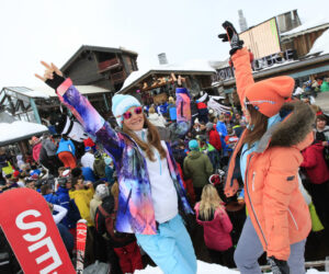 To mennesker i farverige vinterdragter fejrer til en udendørs afterski-fest på deres spændende skirejser, omgivet af en livlig menneskemængde og sneklædte skihytter. En person løfter begge arme, og den anden holder en skistav.