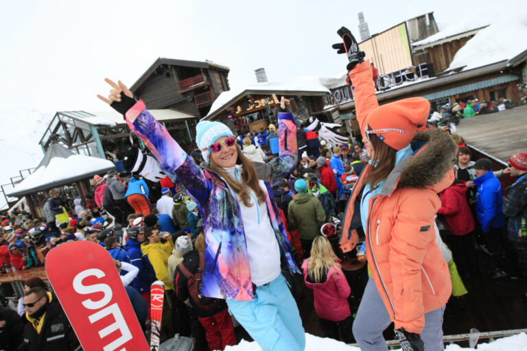 To mennesker i farverige vinterdragter fejrer til en udendørs afterski-fest på deres spændende skirejser, omgivet af en livlig menneskemængde og sneklædte skihytter. En person løfter begge arme, og den anden holder en skistav.