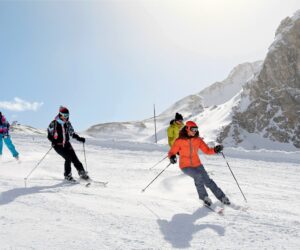 Fire personer på ski ned ad bakke på en sneklædt bjergskråning. De er klædt i farverigt skiudstyr, inklusive hjelme og beskyttelsesbriller. Baggrunden viser en klar blå himmel og en stenet bjergtop.