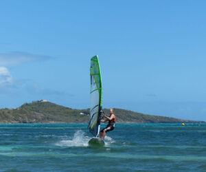 En person, der windsurfer på et turkisblåt hav under en klar blå himmel. Bakker og grønne områder er synlige i baggrunden, med blide bølger, der plasker rundt om windsurferen.