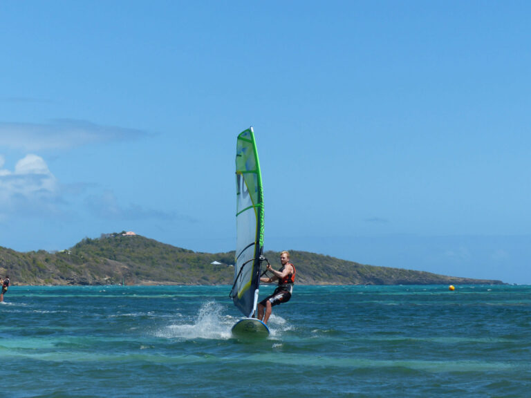 En person, der windsurfer på et turkisblåt hav under en klar blå himmel. Bakker og grønne områder er synlige i baggrunden, med blide bølger, der plasker rundt om windsurferen.