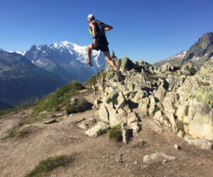 En person i atletisk gear og kasket springer over stenet terræn på en bjergsti. Baggrunden byder på fantastiske bjergtoppe under en klar blå himmel.