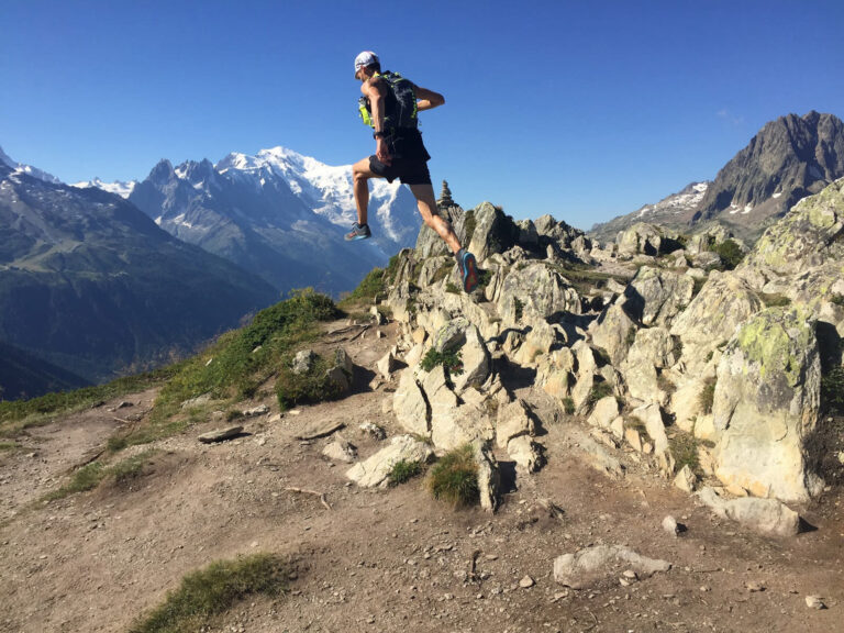 En person i atletisk gear og kasket springer over stenet terræn på en bjergsti. Baggrunden byder på fantastiske bjergtoppe under en klar blå himmel.