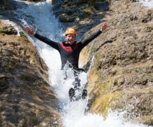 En person iført hjelm og våddragt glider glædeligt ned ad en naturlig vandrutsjebane dannet af sten, med arme løftet og vand sprøjtende rundt. Vejret er solrigt, hvilket fremhæver det omkringliggende stenrige terræn.