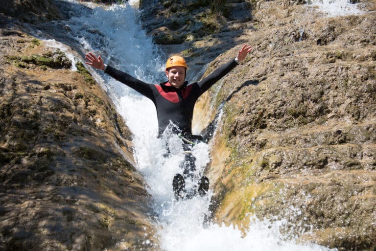 En person iført hjelm og våddragt glider glædeligt ned ad en naturlig vandrutsjebane dannet af sten, med arme løftet og vand sprøjtende rundt. Vejret er solrigt, hvilket fremhæver det omkringliggende stenrige terræn.