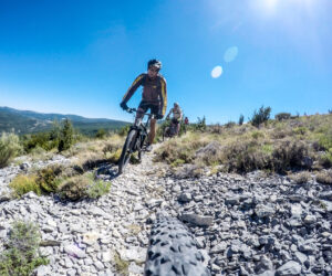 Tre cyklister kører på en stenet bjergsti under en klar blå himmel. Forgrunden viser den forrevne sti og grønne områder på begge sider, hvor sollys skaber linseudstråling. Denne spændende sportsferie fanger eventyrlysten mod et betagende bjerglandskab i baggrunden.