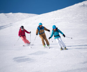 Tre skiløbere i farverige outfits glider ned ad en sneklædt skråning under en klar blå himmel. De bærer hjelme og skibriller og bevæger sig parallelt på en præpareret skiløjpe.