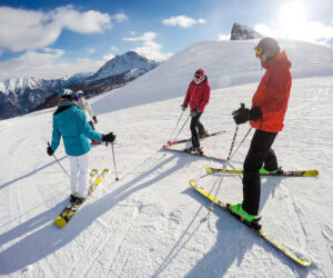 Fire skiløbere i farverigt vinterudstyr står på en sneklædt bjergskråning under en klar blå himmel med majestætiske tinder i baggrunden. Sollys kaster lange skygger, og gruppen ser ud til at tale, inden de går ned ad bakke.