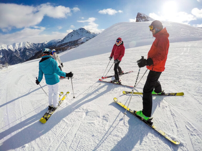 Fire skiløbere i farverigt vinterudstyr står på en sneklædt bjergskråning under en klar blå himmel med majestætiske tinder i baggrunden. Sollys kaster lange skygger, og gruppen ser ud til at tale, inden de går ned ad bakke.