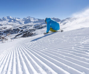 En skiløber i blå jakke og gule bukser karrer ned ad en nypræpareret bakke med snesprøjtning bagved, der minder om en idyllisk skiferie. Majestætiske snedækkede bjerge og en klar blå himmel skaber en fantastisk kulisse.