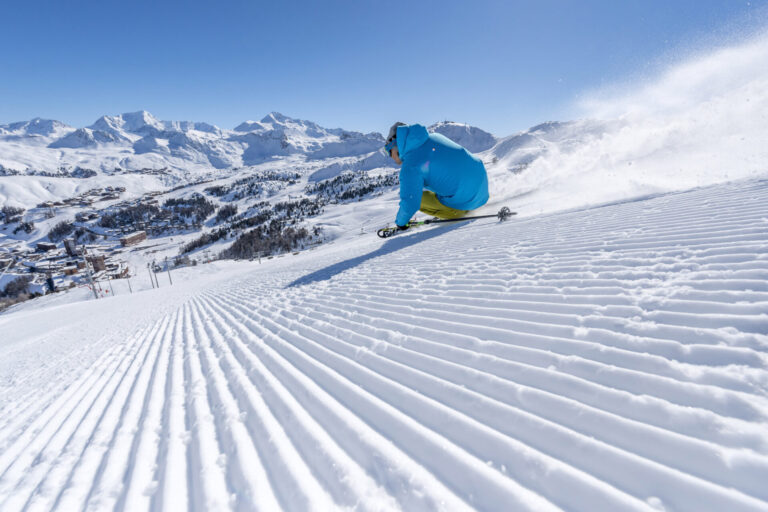 En skiløber i blå jakke og gule bukser karrer ned ad en nypræpareret bakke med snesprøjtning bagved, der minder om en idyllisk skiferie. Majestætiske snedækkede bjerge og en klar blå himmel skaber en fantastisk kulisse.