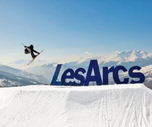 En skiløber udfører et trick i luften over en snedækket rampe med et stort Les Arcs-skilt i baggrunden. Snedækkede bjerge og klar blå himmel fuldender det naturskønne vinterlandskab.