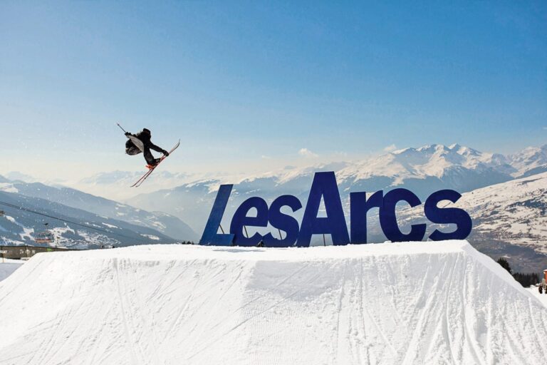 En skiløber udfører et trick i luften over en snedækket rampe med et stort Les Arcs-skilt i baggrunden. Snedækkede bjerge og klar blå himmel fuldender det naturskønne vinterlandskab.