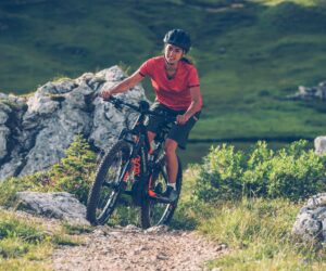 En person i rød skjorte og hjelm kører på mountainbike på en stenet sti omgivet af grønne områder og store sten, der minder om en spændende sportsferie. Scenen formidler en følelse af eventyr og nydelse i naturen.