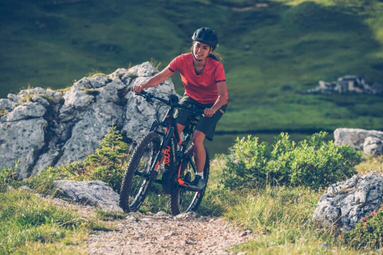 En person i rød skjorte og hjelm kører på mountainbike på en stenet sti omgivet af grønne områder og store sten, der minder om en spændende sportsferie. Scenen formidler en følelse af eventyr og nydelse i naturen.