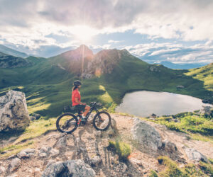 En cyklist i rødt og sort gear holder pause på en bjergsti med udsigt over en naturskøn alpine sø. Solen skinner klart og kaster lange skygger over det grønne landskab og klippetoppe og byder på et perfekt sportsferie øjebliksbillede under en delvist overskyet himmel.