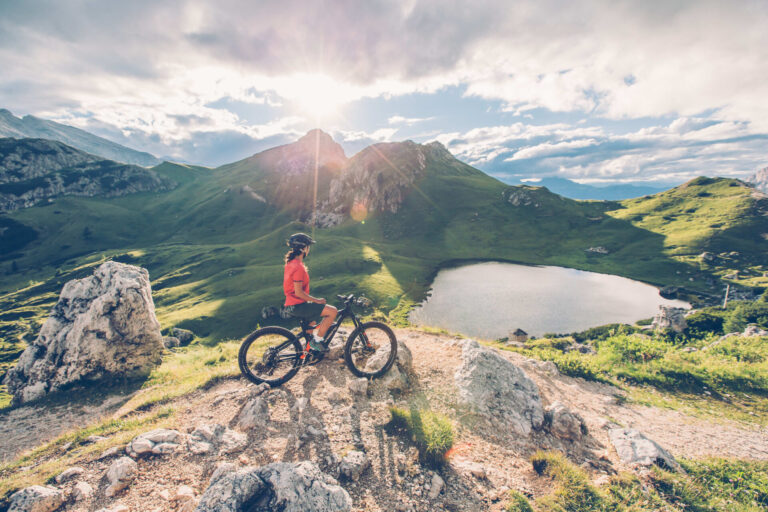 En cyklist i rødt og sort gear holder pause på en bjergsti med udsigt over en naturskøn alpine sø. Solen skinner klart og kaster lange skygger over det grønne landskab og klippetoppe og byder på et perfekt sportsferie øjebliksbillede under en delvist overskyet himmel.