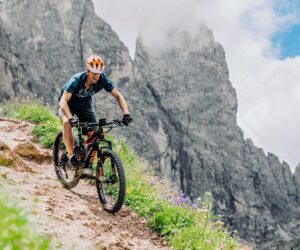 En person i orange hjelm cykler ned ad en stenet sti med stejle klipper i baggrunden. Himlen er delvist overskyet, og vilde blomster er spredt langs stien.