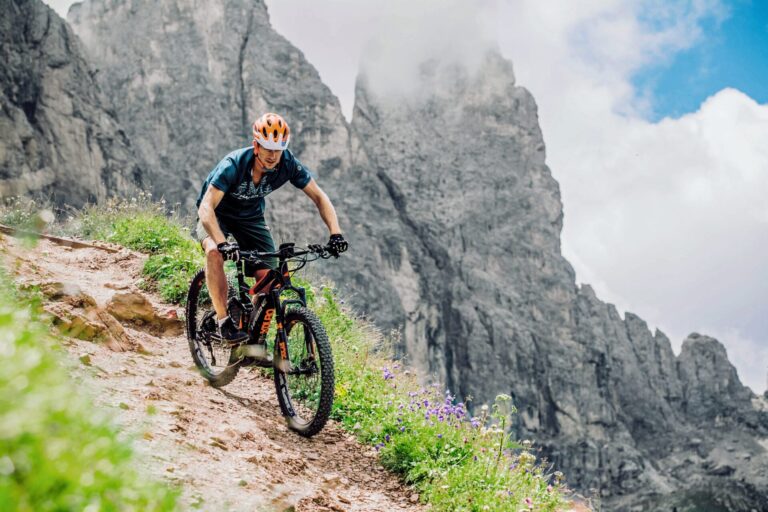 En person i orange hjelm cykler ned ad en stenet sti med stejle klipper i baggrunden. Himlen er delvist overskyet, og vilde blomster er spredt langs stien.