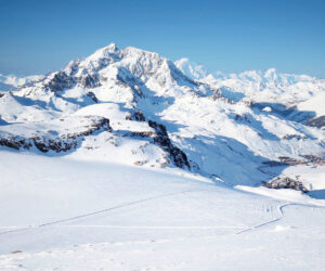 Et betagende vinterlandskab med snedækkede bjerge under en klar blå himmel. Udsigten omfatter forrevne tinder og vidtstrakte, uberørte snemarker, hvilket skaber en fredfyldt og majestætisk atmosfære.