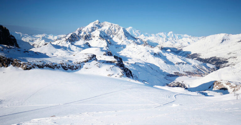 Et betagende vinterlandskab med snedækkede bjerge under en klar blå himmel. Udsigten omfatter forrevne tinder og vidtstrakte, uberørte snemarker, hvilket skaber en fredfyldt og majestætisk atmosfære.