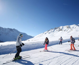 En gruppe mennesker, der står på ski på en sneklædt skråning under en klar blå himmel. Snedækkede bjerge er synlige i baggrunden. Skiløberne er klædt i vinterudstyr, og sollyset kaster lange skygger på sneen.