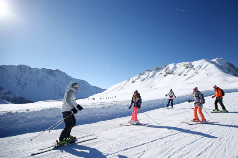 En gruppe mennesker, der står på ski på en sneklædt skråning under en klar blå himmel. Snedækkede bjerge er synlige i baggrunden. Skiløberne er klædt i vinterudstyr, og sollyset kaster lange skygger på sneen.