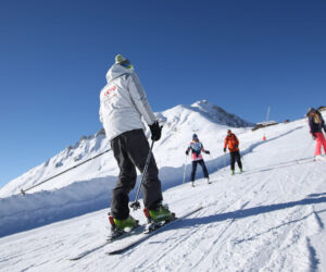 En skiløber i hvid jakke og grøn hjelm kører ned ad en sneklædt bjergskråning, efterfulgt af en gruppe skiløbere. Den klare blå himmel og snedækkede tinder giver en naturskøn kulisse.