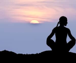 Silhuet af en person, der mediterer i en positur med krydsede ben på en bakketop under solnedgang. Solen er delvist synlig og kaster et varmt skær hen over den fredfyldte himmel.