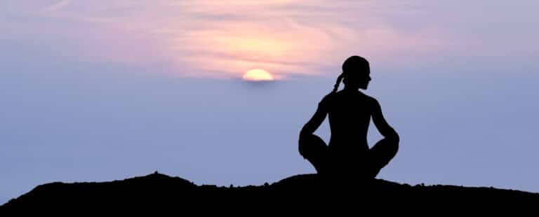 Silhuet af en person, der mediterer i en positur med krydsede ben på en bakketop under solnedgang. Solen er delvist synlig og kaster et varmt skær hen over den fredfyldte himmel.