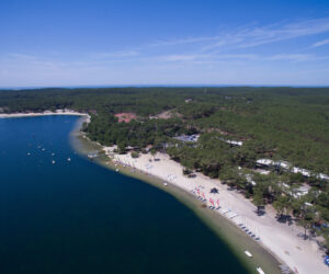 Luftfoto af en sandstrand med klart blåt vand, oversået med små både. Kystlinjen buer blidt, omkranset af tæt grøn skov. Farvede flag og strandfaciliteter er synlige, og himlen er klar med et par tjavsede skyer.