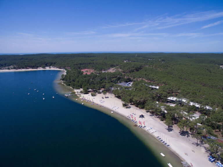 Luftfoto af en sandstrand med klart blåt vand, oversået med små både. Kystlinjen buer blidt, omkranset af tæt grøn skov. Farvede flag og strandfaciliteter er synlige, og himlen er klar med et par tjavsede skyer.