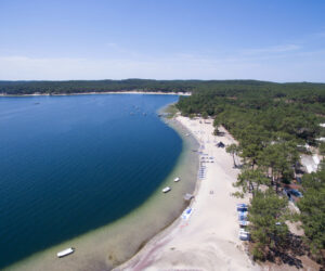Luftfoto af en fredfyldt strand med hvidt sand, der buer langs en dybblå sø. Små både ligger for anker nær kysten, omgivet af en frodig grøn skov under en klar blå himmel.