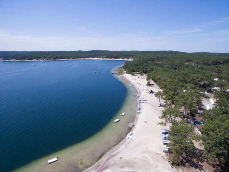 Luftfoto af en fredfyldt strand med hvidt sand, der buer langs en dybblå sø. Små både ligger for anker nær kysten, omgivet af en frodig grøn skov under en klar blå himmel.