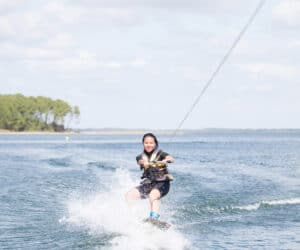 En person, der wakeboarder på en solskinsdag og sprøjter vand, mens de holder fast i et reb, der er fastgjort til en båd. Vandet er klart med blide bølger, og en række træer er synlige i baggrunden under en delvist overskyet himmel.