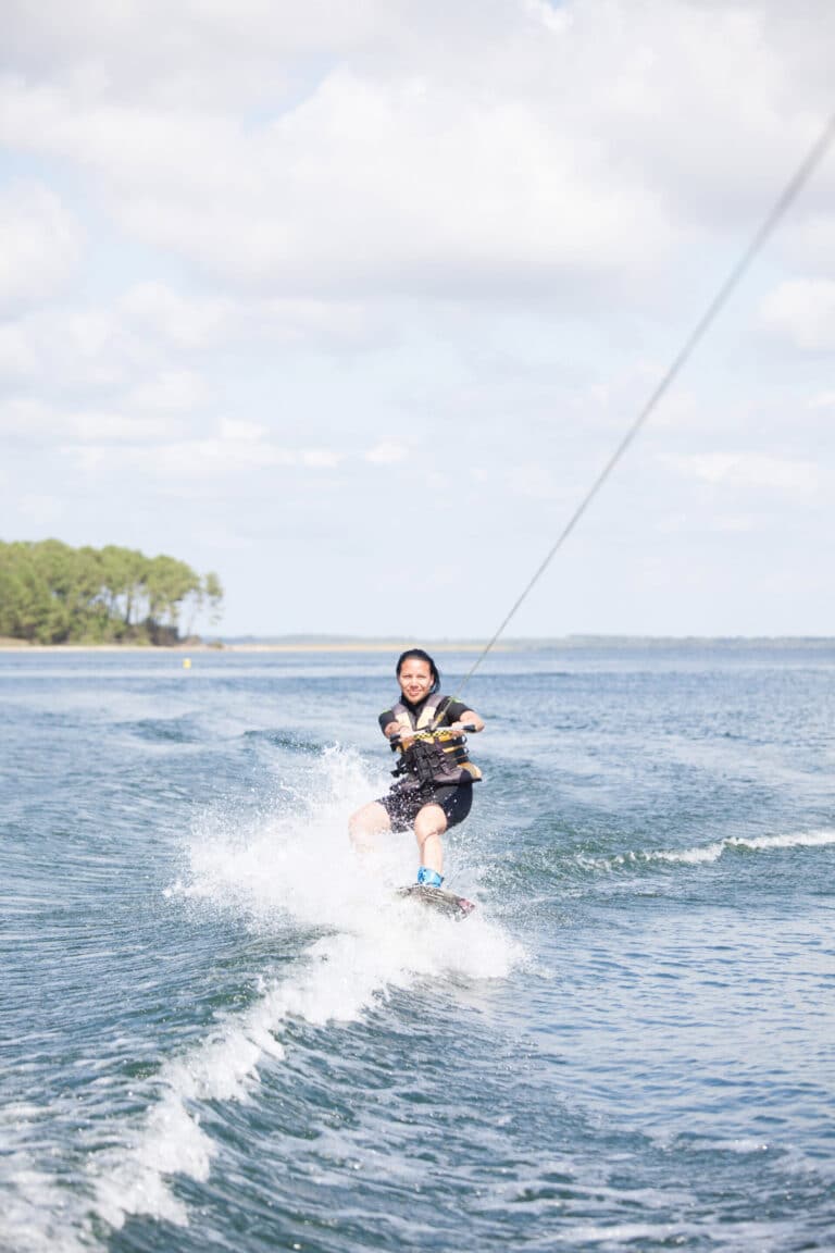 En person, der wakeboarder på en solskinsdag og sprøjter vand, mens de holder fast i et reb, der er fastgjort til en båd. Vandet er klart med blide bølger, og en række træer er synlige i baggrunden under en delvist overskyet himmel.