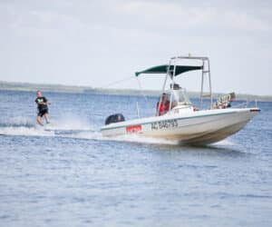 En person wakeboarder på vandet bag en motorbåd med en grøn baldakin. Båden bevæger sig med fart og skaber bølger, og wakeboarderen holder balancen på bølgerne. Baggrunden viser en klar himmel og en fjern kystlinje.